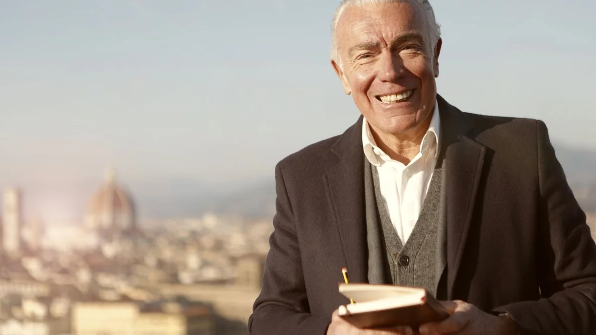 An older man smiling confidently outdoors with a sunlit city skyline behind him